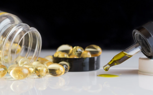 Clear capsules spilling from a transparent bottle next to a dropper releasing a drop of yellow oil on a white surface with black background.