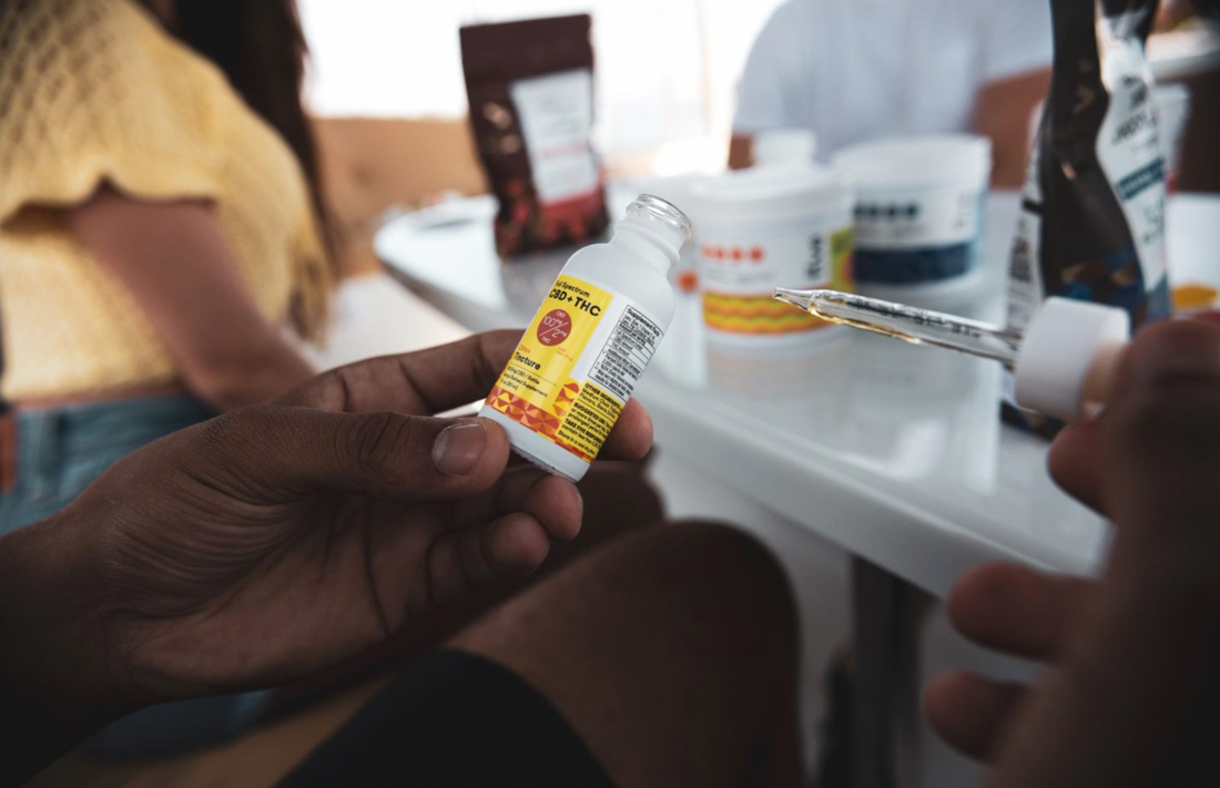 Hand holding a CBD + THC tincture bottle with a dropper near a table with other containers