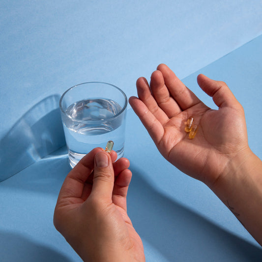Two hands holding three translucent capsules near a glass of water on a blue surface.