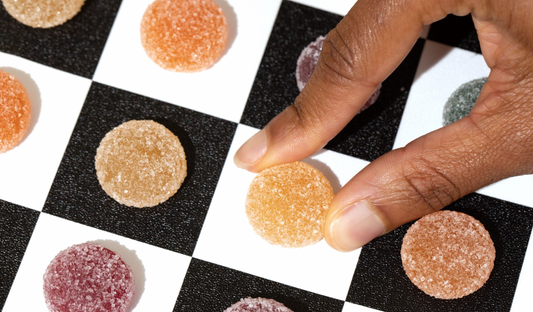 A hand picking an orange sugar-coated gummy candy from a black and white checkerboard surface with other gummies in orange, beige, purple, and green colors.