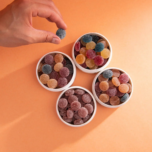 Top-down view: hand holding a blue sugar-coated gummy above four white tubs of assorted sugar-coated gummies on orange