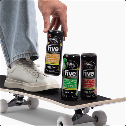 Hand placing a 'five' THC can on a skateboard deck near three other 'five' cans; labels read five, FAST-ACTING, 100MG THC.