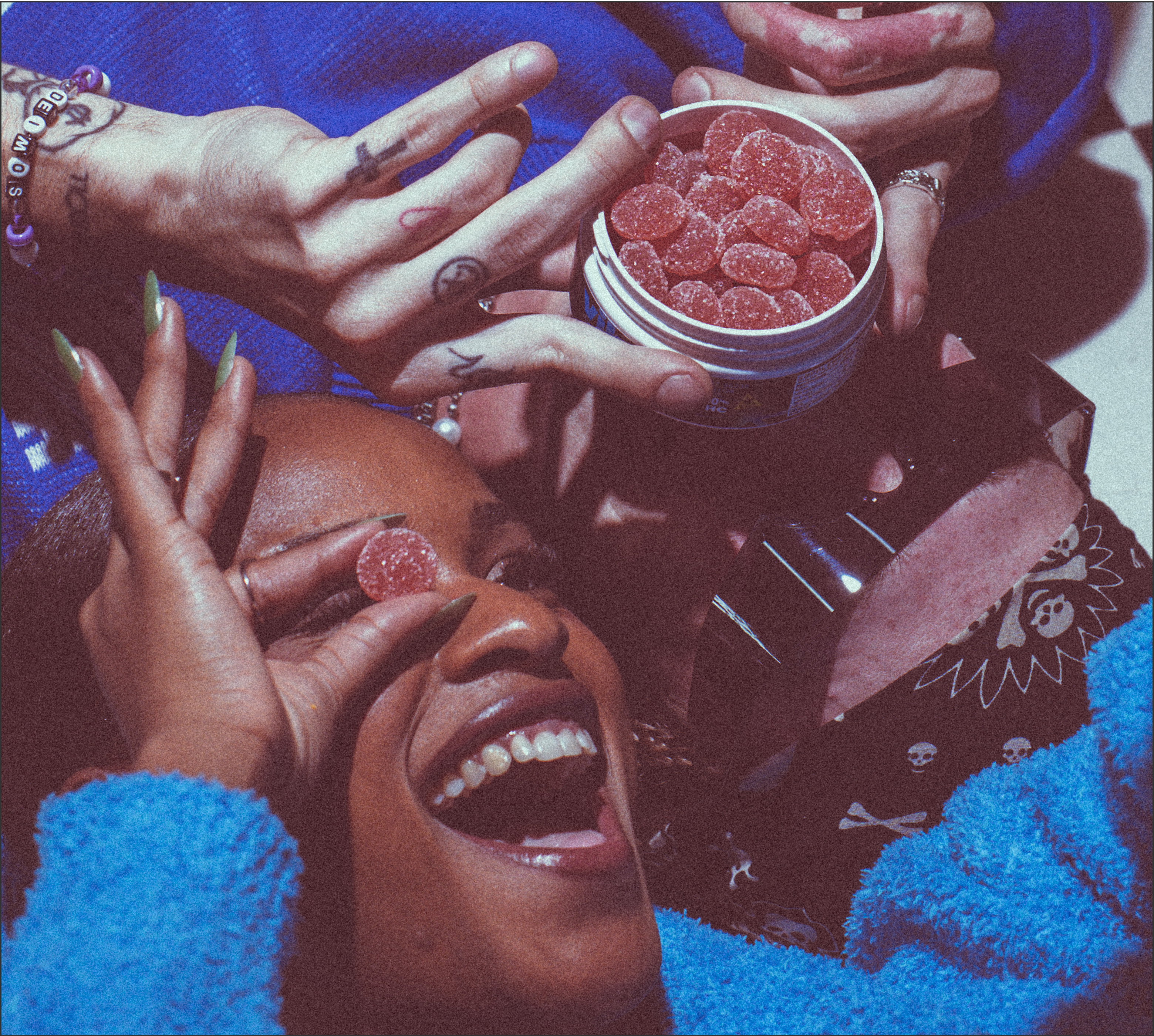 Hands holding a jar of red sugar-coated gummies above a laughing person holding one gummy to their eye; beaded bracelet reads DELMOS.