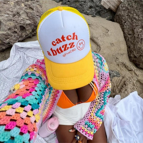 Person wearing a yellow and white cap with red text "catch a buzz @FiveCBD" and a colorful crochet cardigan, sitting on a sandy surface with rocks nearby.