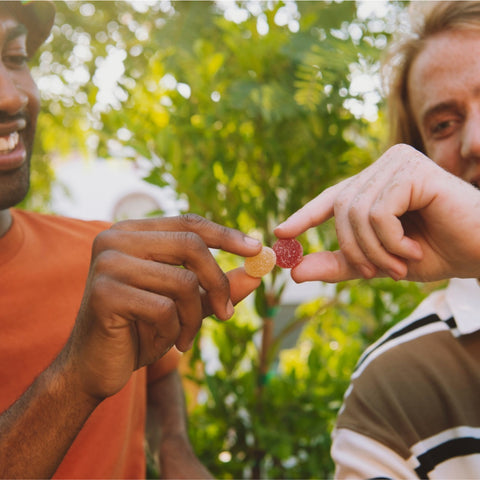 Two people holding and touching small round gummy candies outdoors with greenery in the background.
