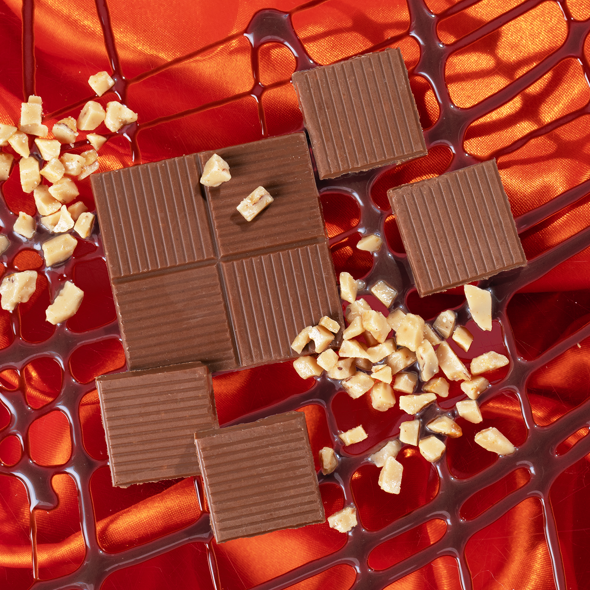 Milk chocolate squares with ridged tops, chocolate syrup lattice and chopped nuts on red satin fabric.