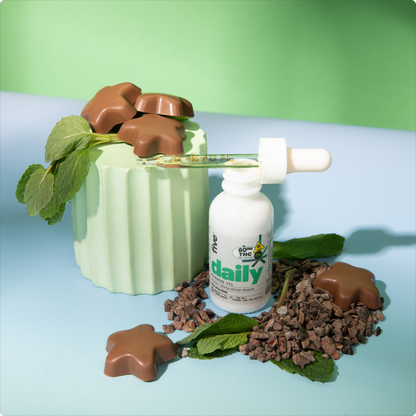 CBD oil bottle labeled 'daily' with dropper, surrounded by star-shaped chocolates, mint leaves, and cacao nibs on a pastel background.