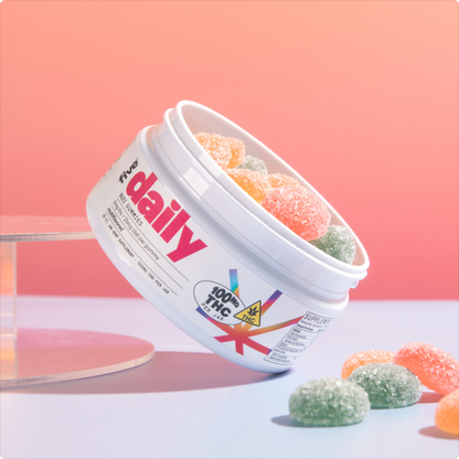 White tub labeled "daily" and "100% THC" tipped on its side with orange and green sugar‑coated gummy candies on a pink backdrop.