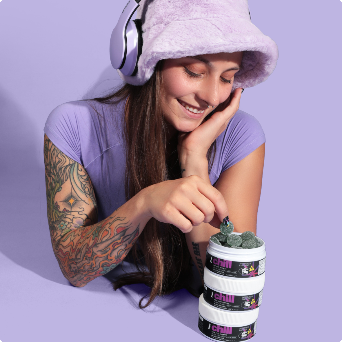 Smiling woman in purple hat and headphones picking a gummy from stacked jars labeled chill THC 200