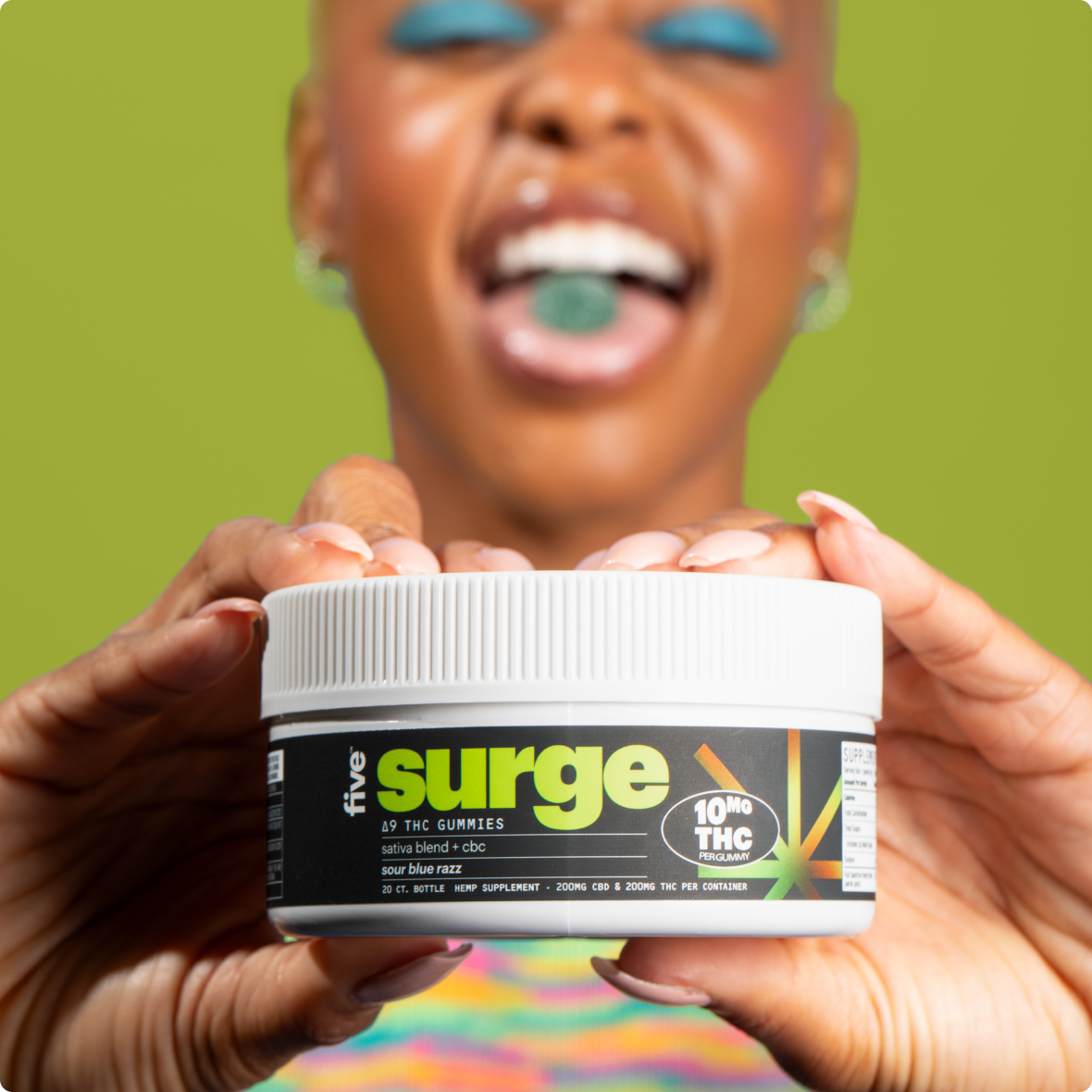 Person holding jar labeled five surge with 10MG THC PER GUMMY, green gummy on their tongue, blue eye makeup, lime background