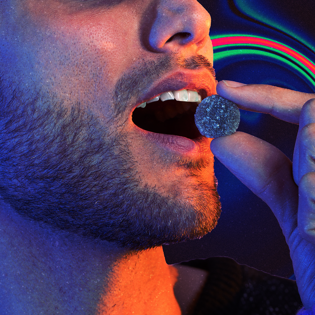 Close-up of a bearded man's open mouth as fingers hold a sugar-coated gummy near his lips, neon lighting.