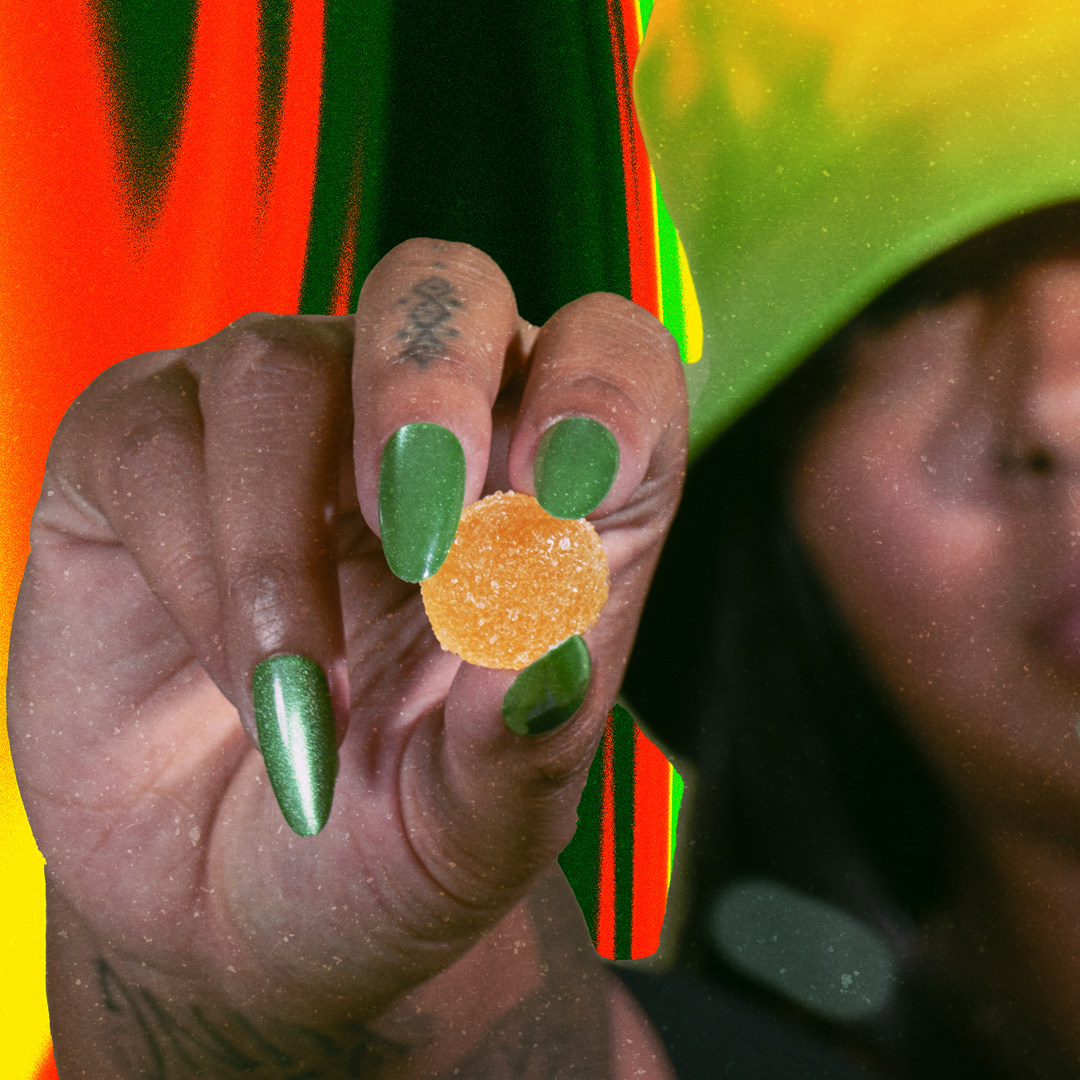 Hand with green metallic nails and small finger tattoo holding a sugar-coated orange gummy.