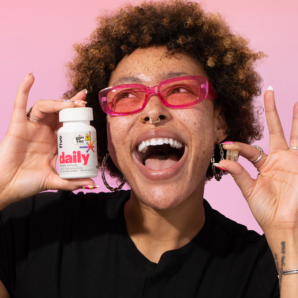 Person smiling, wearing pink sunglasses, holds white bottle labeled 'five daily 60MG THC MICRODOSE CAPSULES' while holding a capsule.