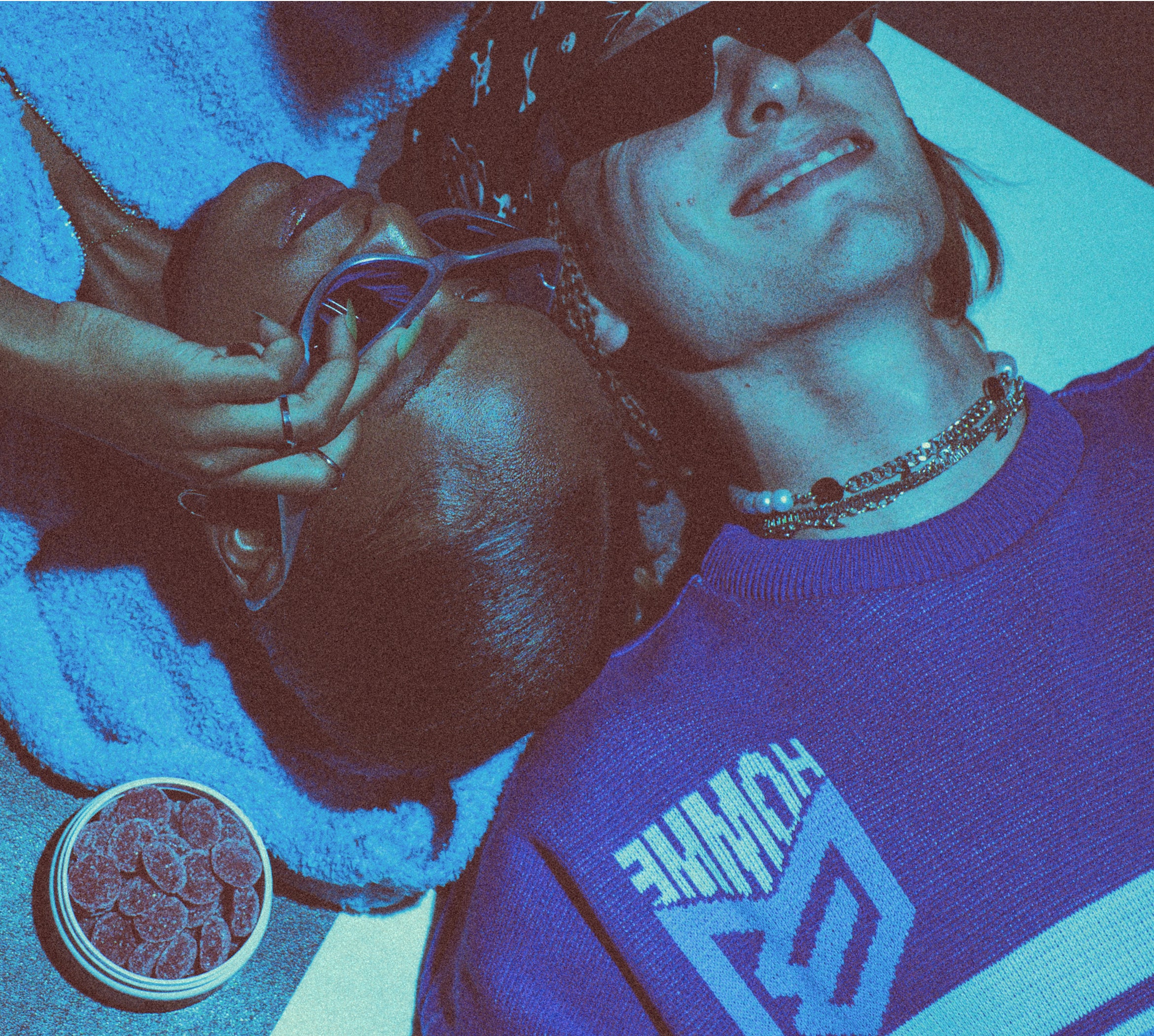 Two people lying down close together, one wearing blue sunglasses and a blue fuzzy jacket, the other wearing a patterned hat, necklace, and a blue sweater with the word 'WHMME' visible. A container with round objects is near them.