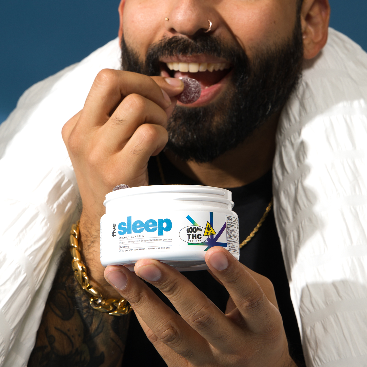 Bearded person holding jar labeled five sleep KNOCKOUT GUMMIES blackberry 100mg THC per jar SUPPLEMENT, putting a gummy in their mouth.