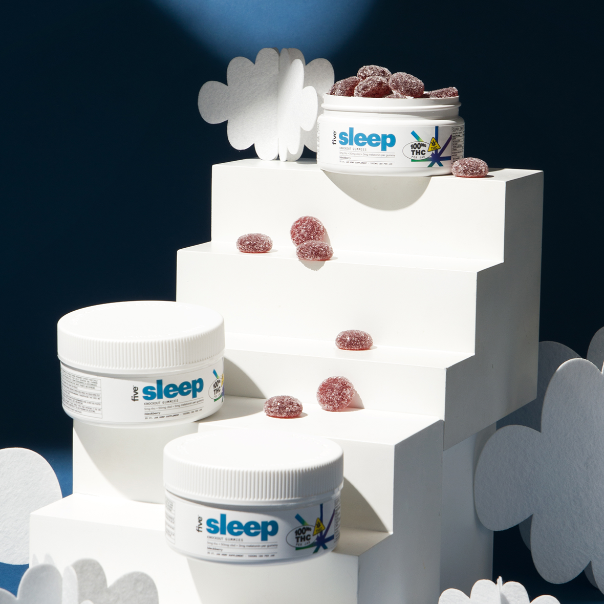 Three white jars labeled five sleep and 100MG THC on white stepped display, with sugar-coated red gummy candies scattered.
