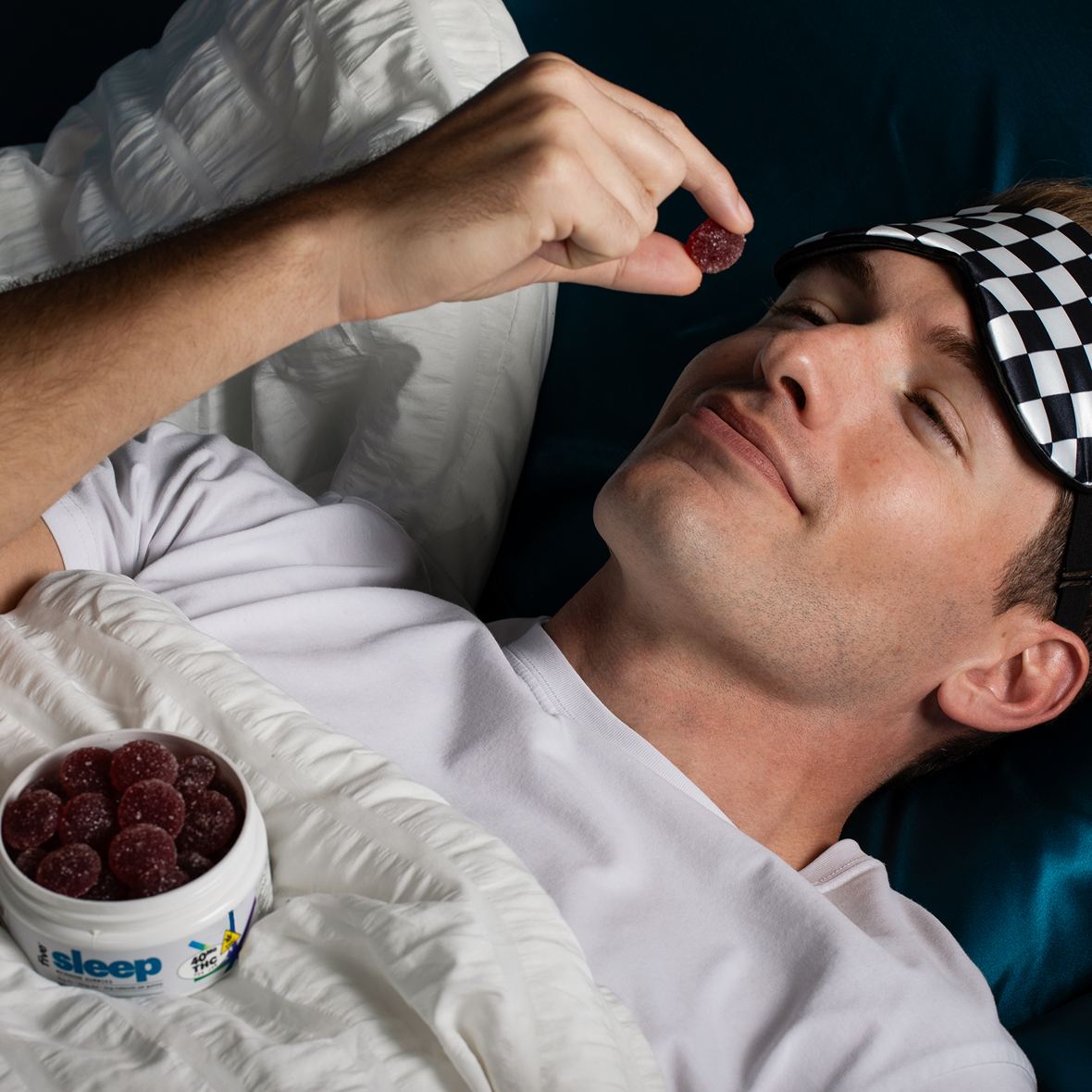 Man lying in bed with a checkered sleep mask, holding a red gummy; tub on blanket labeled 'sleep' and '40 THC'.